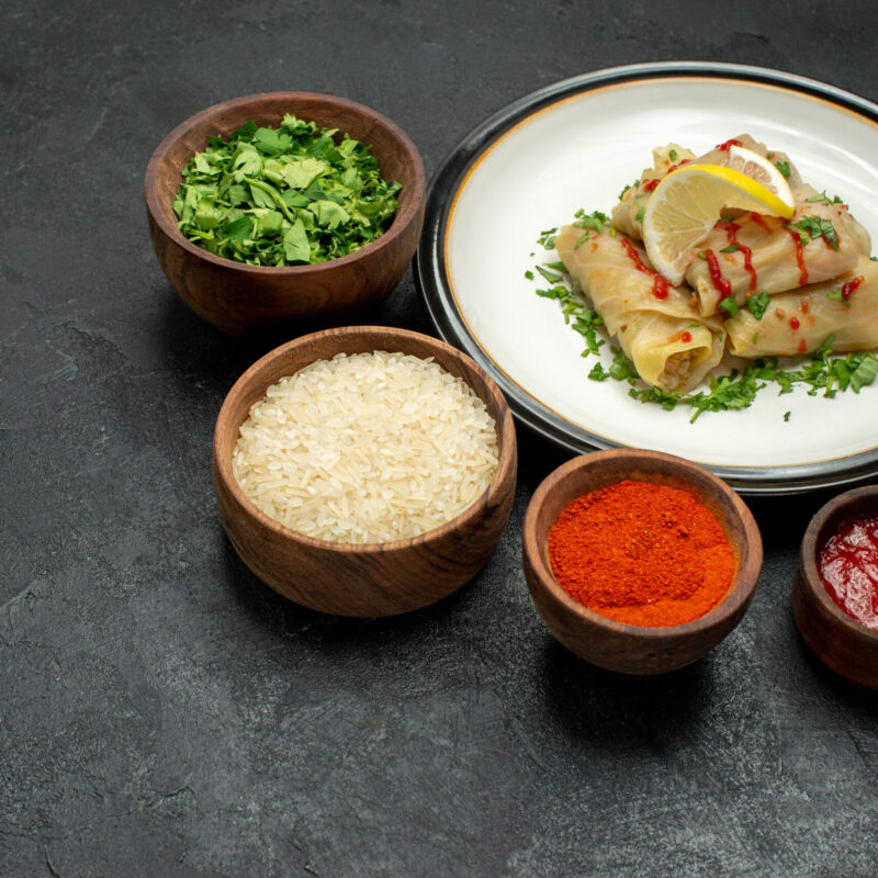 side-close-up-view-food-white-plate-plate-stuffed-cabbage-with-lemon-herbs-sauce-bowls-spices-rice-herbs-sauce-dark-background
