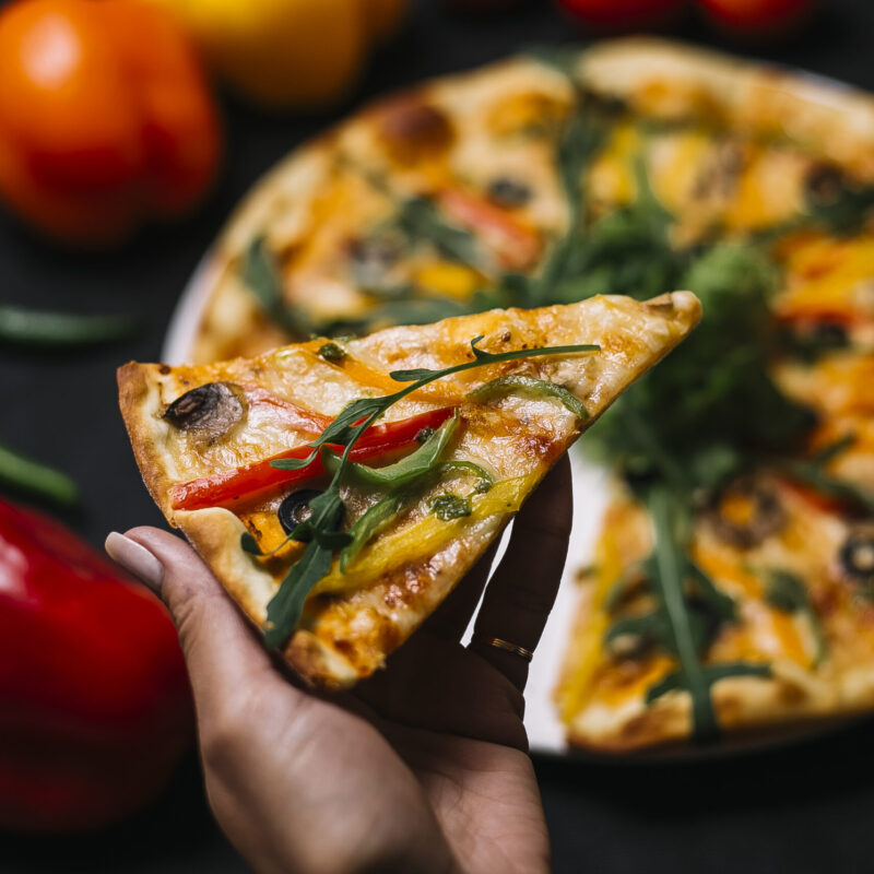 side-view-hand-holding-slice-italian-pizza-with-colorful-bell-peppers-mushrooms-black-olives-urugula-cheese