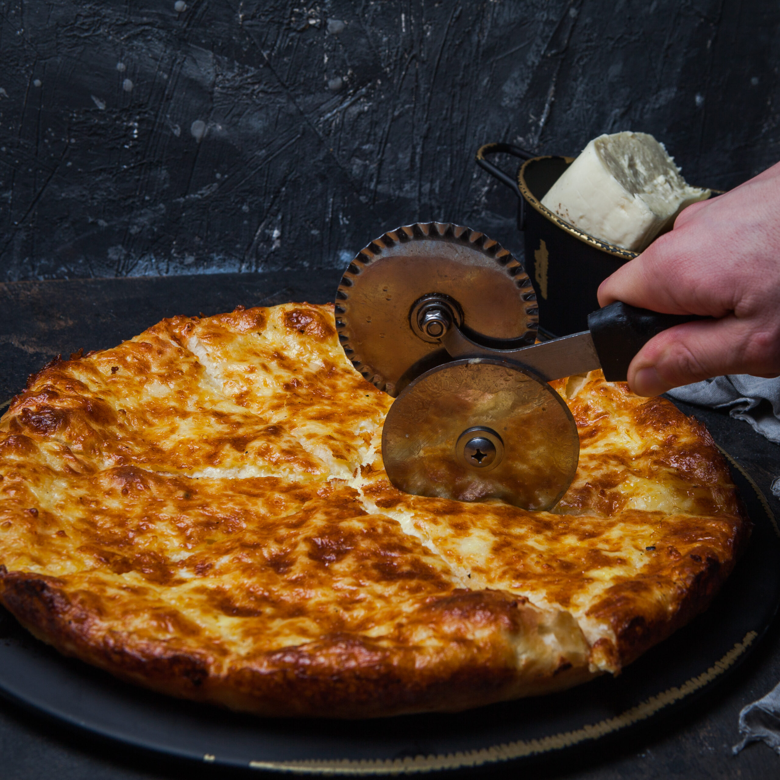 Side view khachapuri with sulguni cheese and pizza cutting knife and human hand in dark plate on stone background