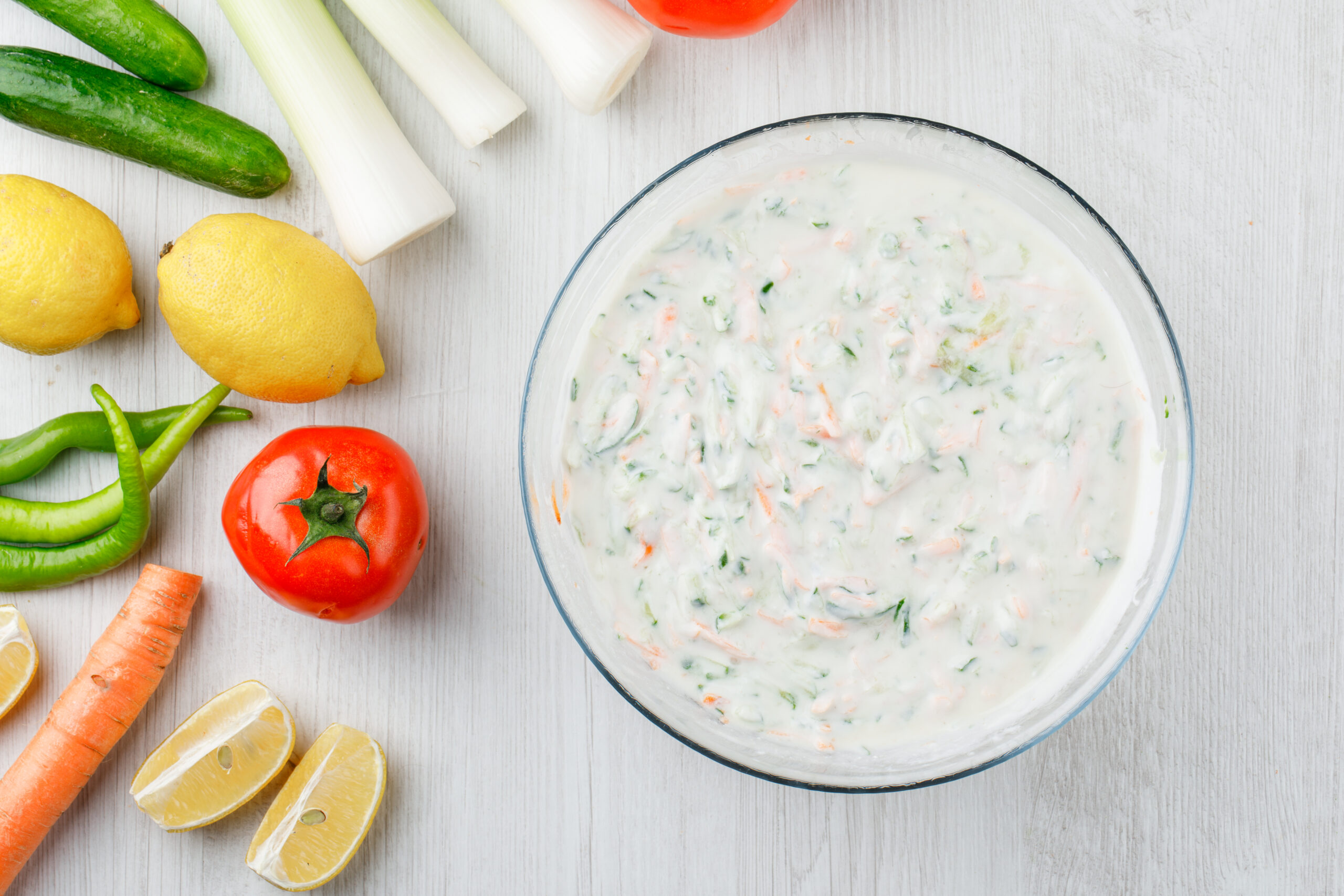 Yogurt salad in a bowl with vegetables and lemons flat lay on a white wooden background